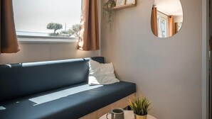 Uma sala de estar moderna com um sofá azul escuro, uma mesa de centro redonda de madeira com uma caneca e uma planta, um espelho redondo na parede, e vista para o exterior através de uma janela com cortinas marrons.