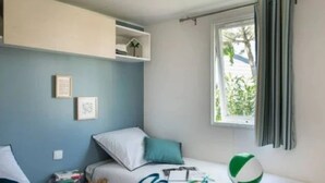A bedroom with a bed, a desk, a window with curtains, and a view of greenery outside.