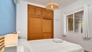 Ein Schlafzimmer mit Bett, Nachttisch, Lampe, Holzschrank, Fenster mit Vorhängen und einer Pendelleuchte.