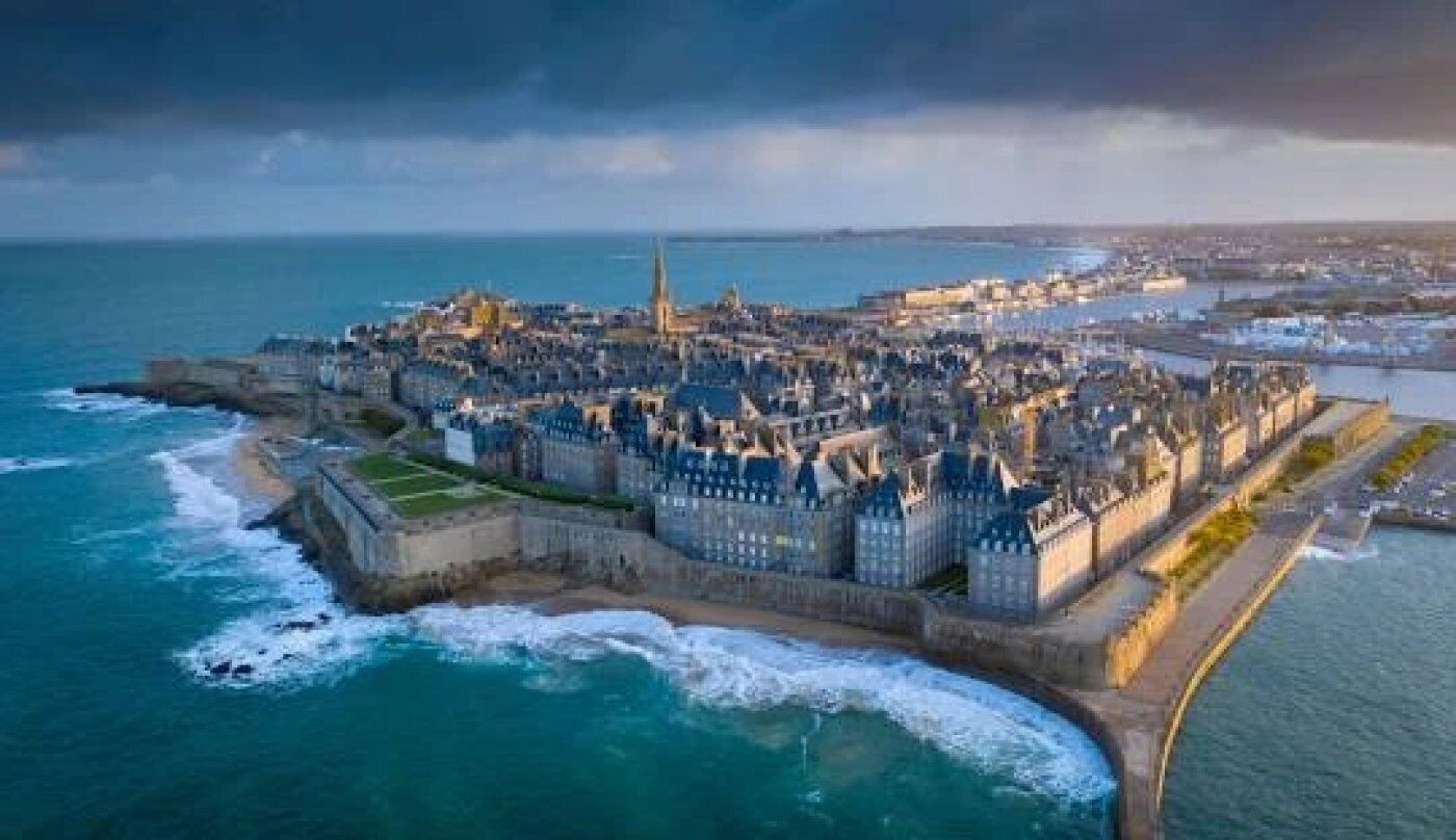 An aerial view of a coastal city with historic architecture, a prominent church, and a fortified harbor.