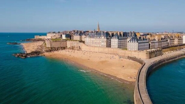 Vista aérea de una ciudad costera con una imponente estructura tipo castillo, una playa de arena y un mar azul cristalino.