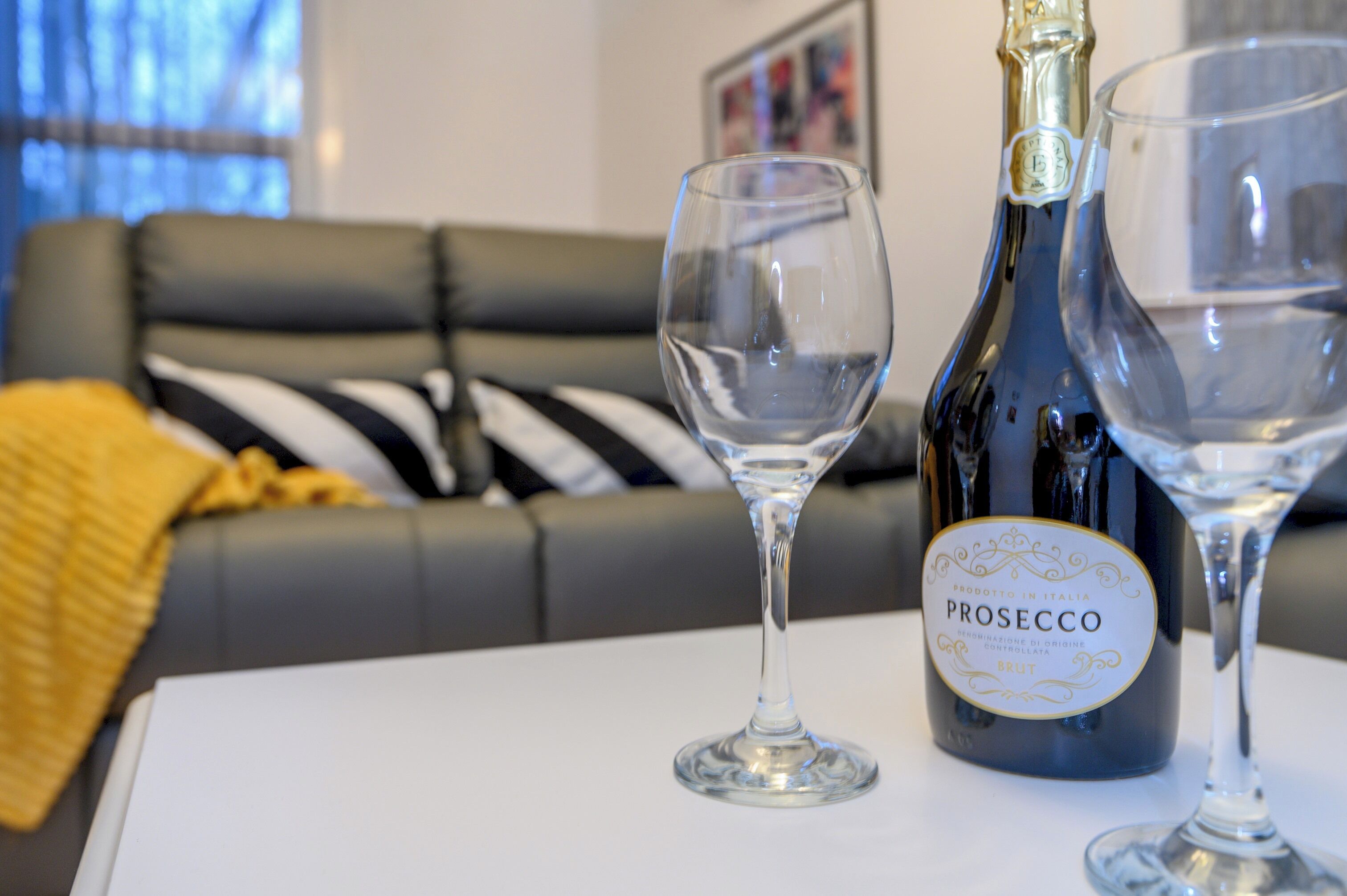 A bottle of Prosecco and two glasses on a table in a room with a black and white striped couch and a yellow throw blanket.