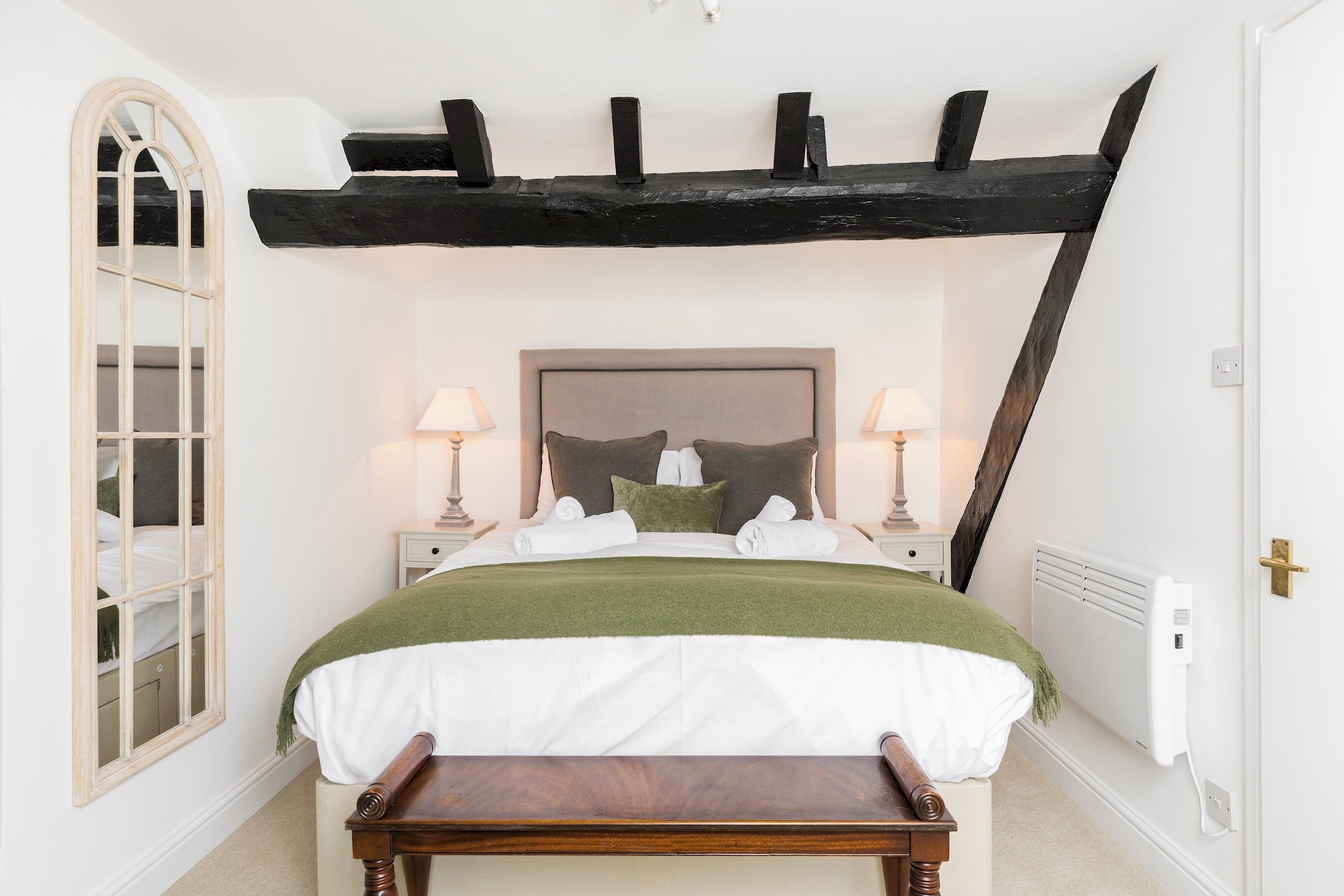 Une chambre à coucher comprenant un lit, des tables de chevet, un banc, un miroir et un radiateur.