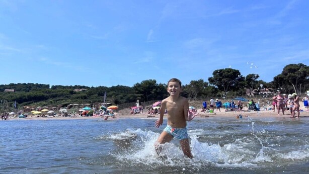 A beach scene with a child playing in the water, umbrellas, and people on the sand. - ZEcamping 3* 200m plage locatif TV clim 28m2 6 pers (Hyères)
