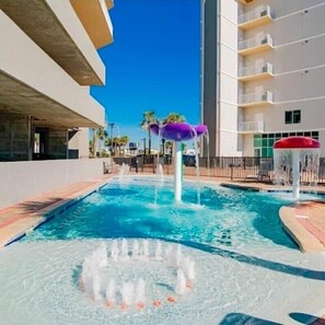 A swimming pool with water fountains, a white building with balconies, and a clear blue sky. - 1704 Seawind w Free Umbrella Svc Double Balcony (Gulf Shores)