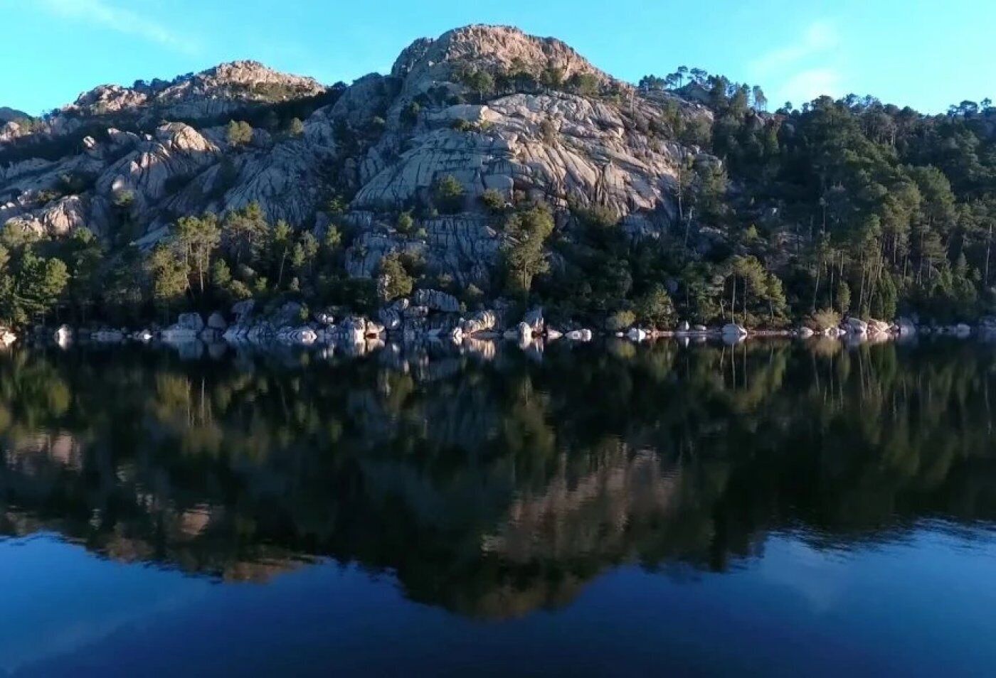 Ein ruhiger See mit einem felsigen Berg im Hintergrund und einigen Gebäuden am Ufer.