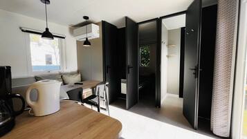 Une pièce moderne et bien éclairée, avec une table à manger en bois, une machine à café et un luminaire suspendu noir. Une porte coulissante donne accès à une salle de bain et à un placard doté d’un rideau à motifs.