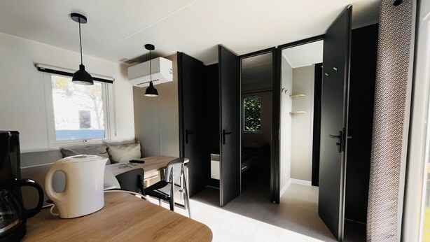 Une pièce moderne et bien éclairée, avec une table à manger en bois, une machine à café et un luminaire suspendu noir. Une porte coulissante donne accès à une salle de bain et à un placard doté d’un rideau à motifs.