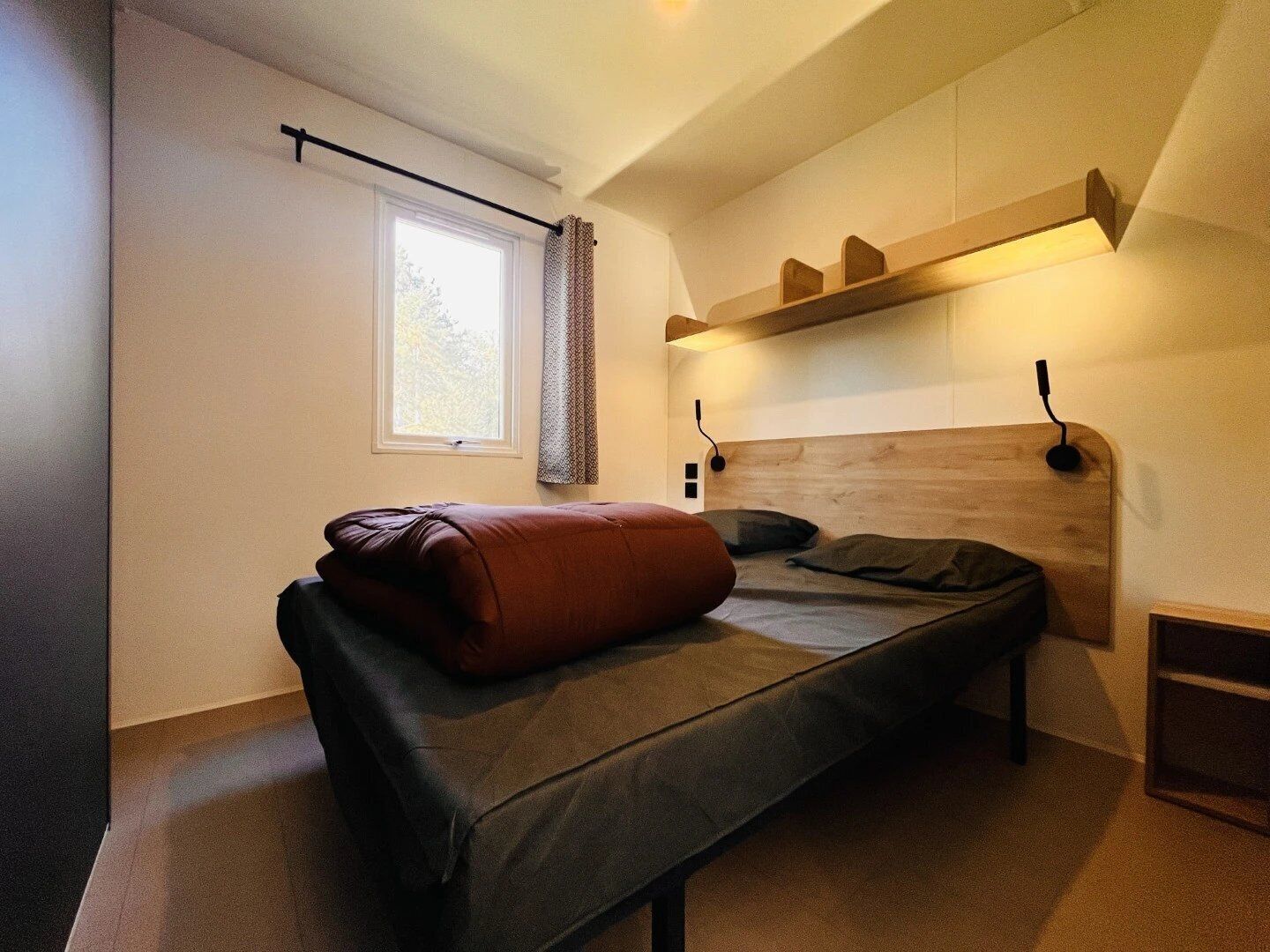 Une petite chambre à coucher moderne, avec un lit simple, une fenêtre avec des rideaux, une étagère au-dessus du lit et une tête de lit dotée d’un éclairage intégré.
