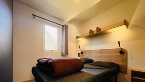 Une petite chambre à coucher moderne, avec un lit simple, une fenêtre avec des rideaux, une étagère au-dessus du lit et une tête de lit dotée d’un éclairage intégré.