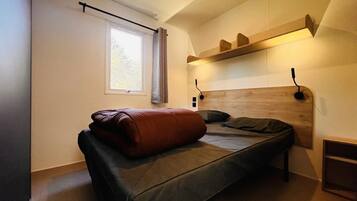 Une petite chambre à coucher moderne, avec un lit simple, une fenêtre avec des rideaux, une étagère au-dessus du lit et une tête de lit dotée d’un éclairage intégré.
