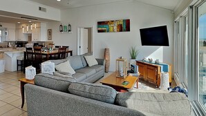 Una sala moderna con un sofá gris, una mesa de centro de madera y un televisor de pantalla plana. Se aprecia un comedor con mesa y sillas, y una cocina al fondo.