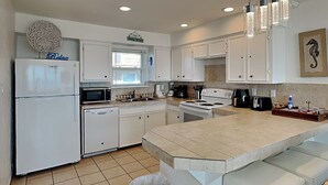 Una cocina moderna con gabinetes blancos, un refrigerador de acero inoxidable, un microondas, una estufa y un fregadero. Hay un bote de basura y una zona de bar con taburetes.