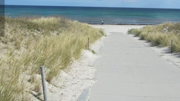 Ein Weg führt zu einem Strand, in der Ferne ist eine Person zu sehen.