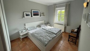 Ein Schlafzimmer mit einem großen Bett, einem Nachttisch, einer Fenster mit Vorhängen und einem Wandregal mit Bildern.