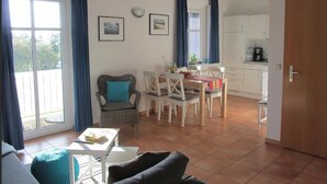 Ein Wohnzimmer mit Sofa, einem Esstisch mit Stühlen, einer Küchenzeile und einem Balkon mit Vorhängen.