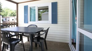 A covered patio with a table and chairs, a window with blue shutters, and a view of a house in the background. - ZEcamping 3*chiens acceptés piscine en bord de rivière avec Canoë (Mazères)