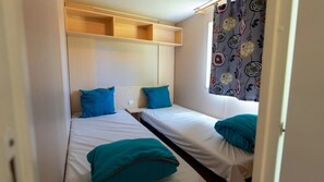 Une petite chambre compacte avec deux lits simples, chacun étant équipé d’un oreiller et d’une couverture. Il y a une fenêtre avec un rideau à motifs floraux, et une étagère au-dessus des lits.