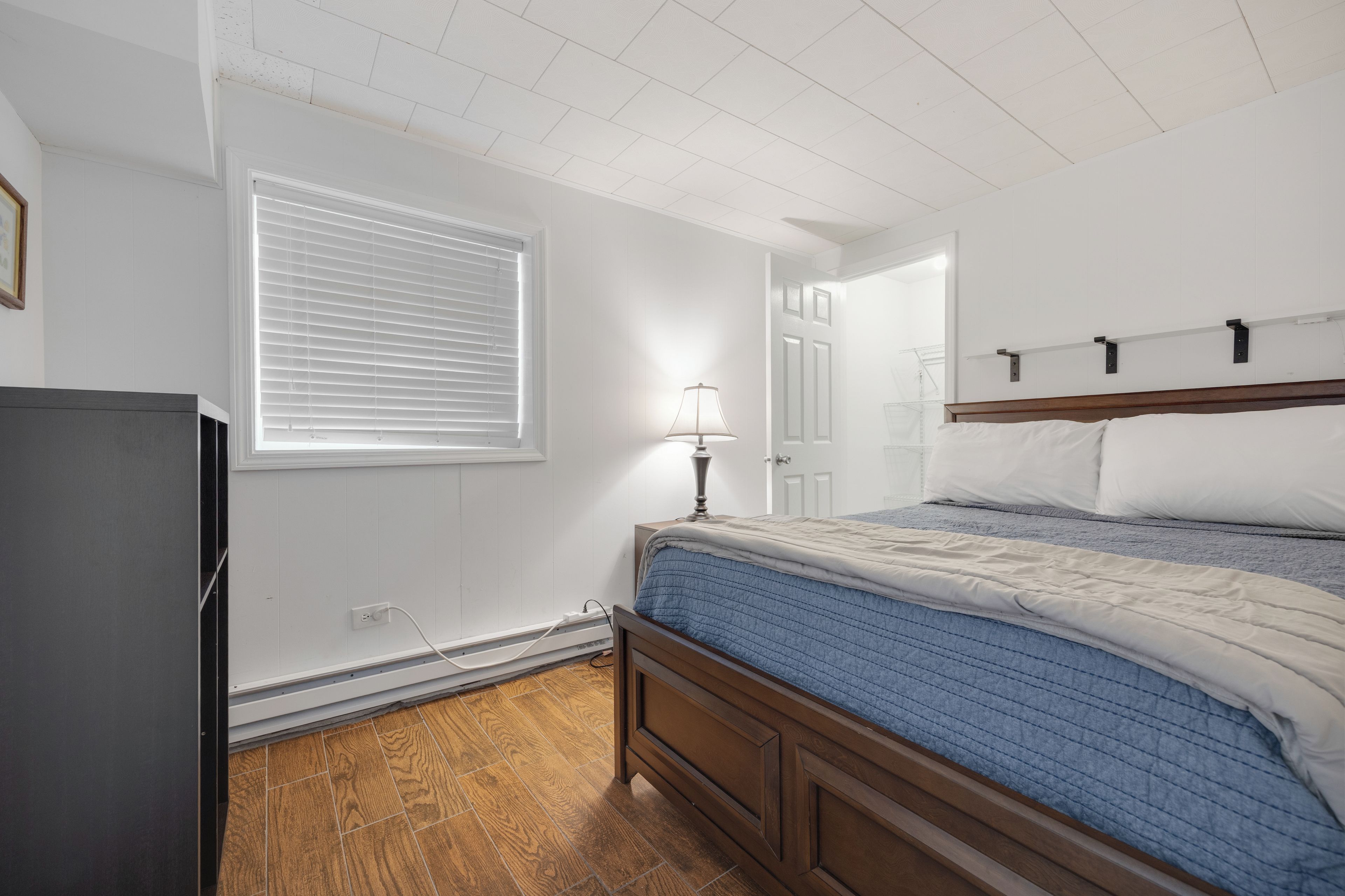 A bedroom with a bed, a bedside table with a lamp, a window with blinds, a door, and a wall-mounted shelf.