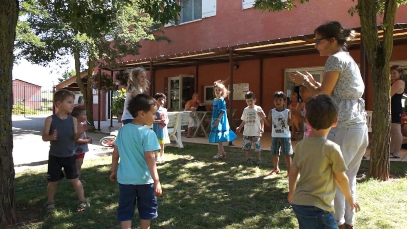 Un groupe d’enfants et d’adultes se trouve à l’extérieur, peut-être dans un parc ou dans la cour d’une école, avec un bâtiment en arrière-plan.