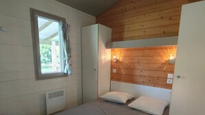 Una habitación pequeña con una cama individual, techo de madera, ventana con cortinas, mesita de noche, armario y una lámpara fijada a la pared.