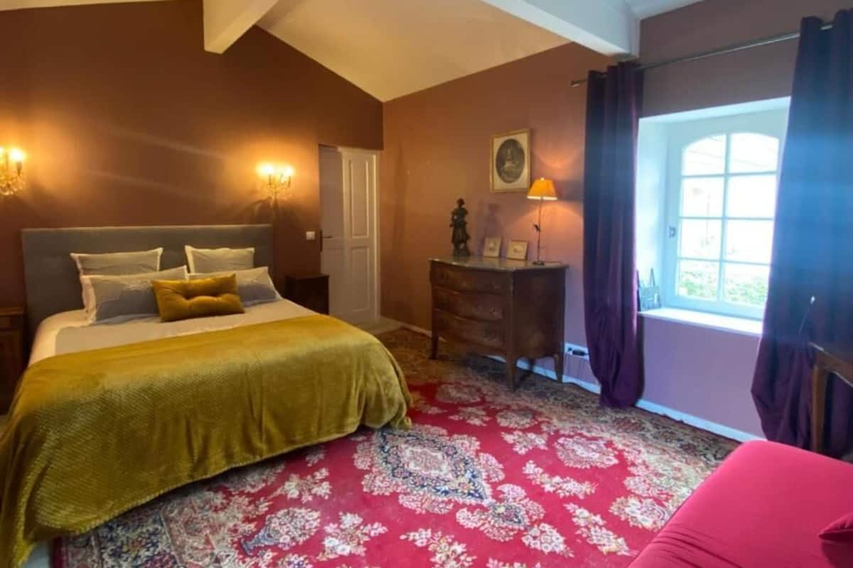 Ein Schlafzimmer mit Bett, Kommode, Lampe, Fenster mit Vorhängen und einer Tür.