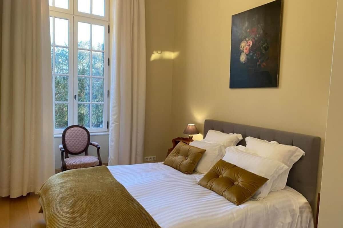 Ein ordentlich bezogenes Bett mit weißer und brauner Bettwäsche, ein Sessel, ein Bild an der Wand und ein Fenster mit Vorhängen.