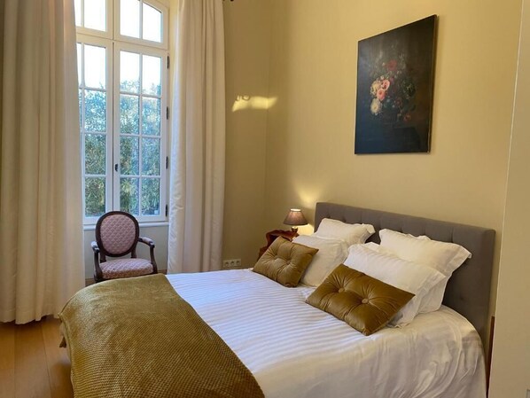 Una cama bien tendida con ropa de cama blanca y marrón, una silla, un cuadro en la pared y una ventana con cortinas.