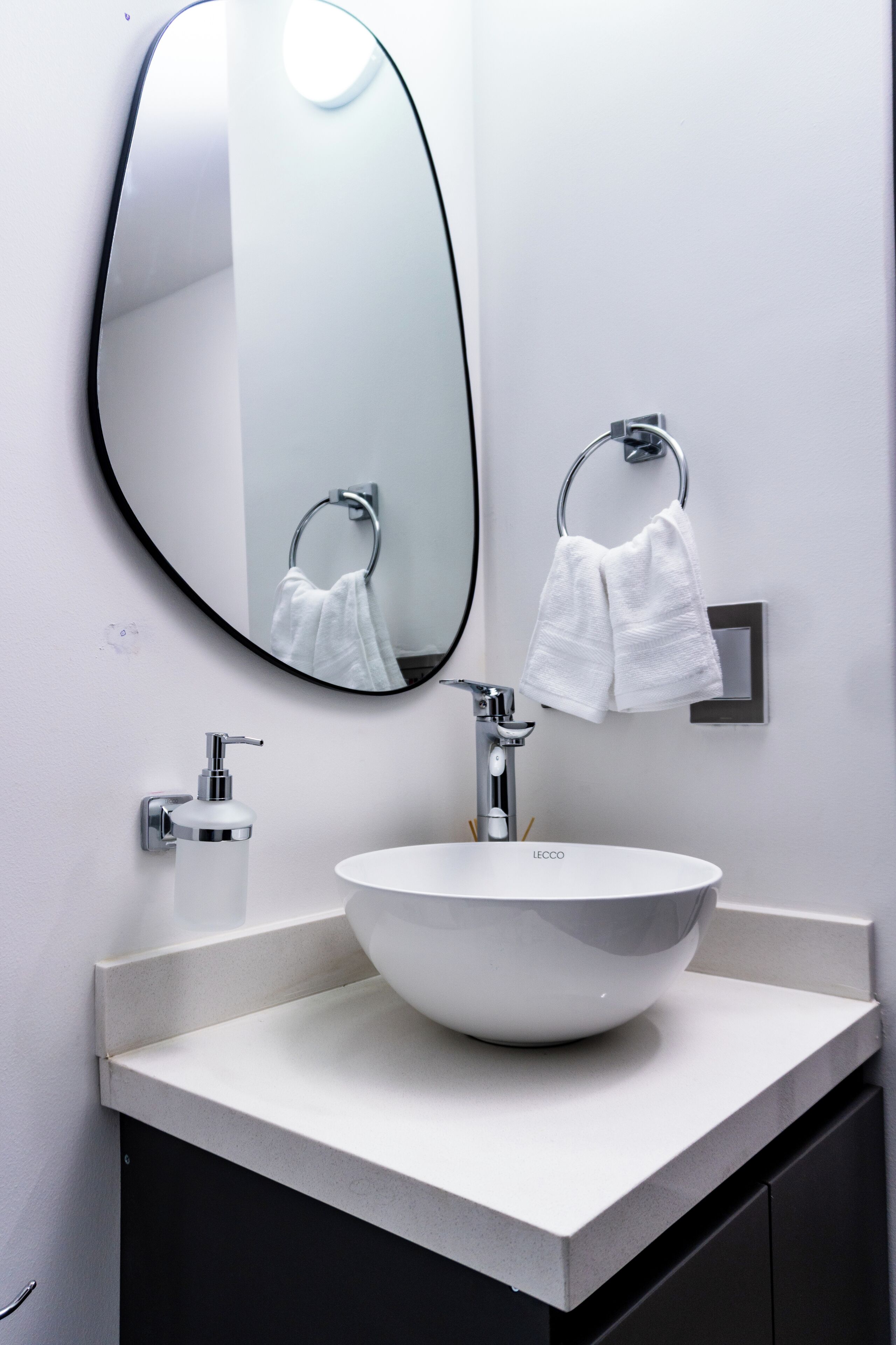 A modern bathroom with a round white sink, a large mirror, and two towels hanging on towel rings.