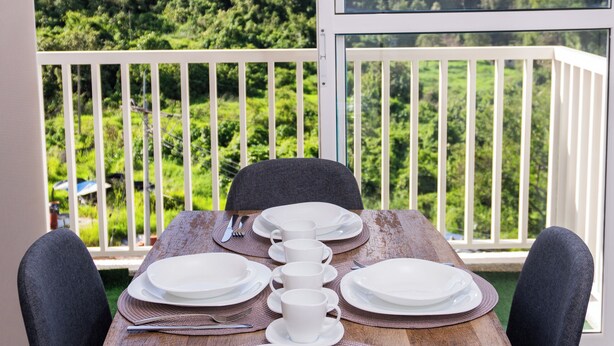 Mesa de jantar posta para quatro, com pratos brancos, talheres e uma xícara, com vista para uma paisagem verde exuberante e um guarda-corpo de varanda branco.
