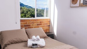 Cama bem arrumada com colcha marrom, um par de toalhas enroladas e um conjunto de artigos de higiene pessoal.