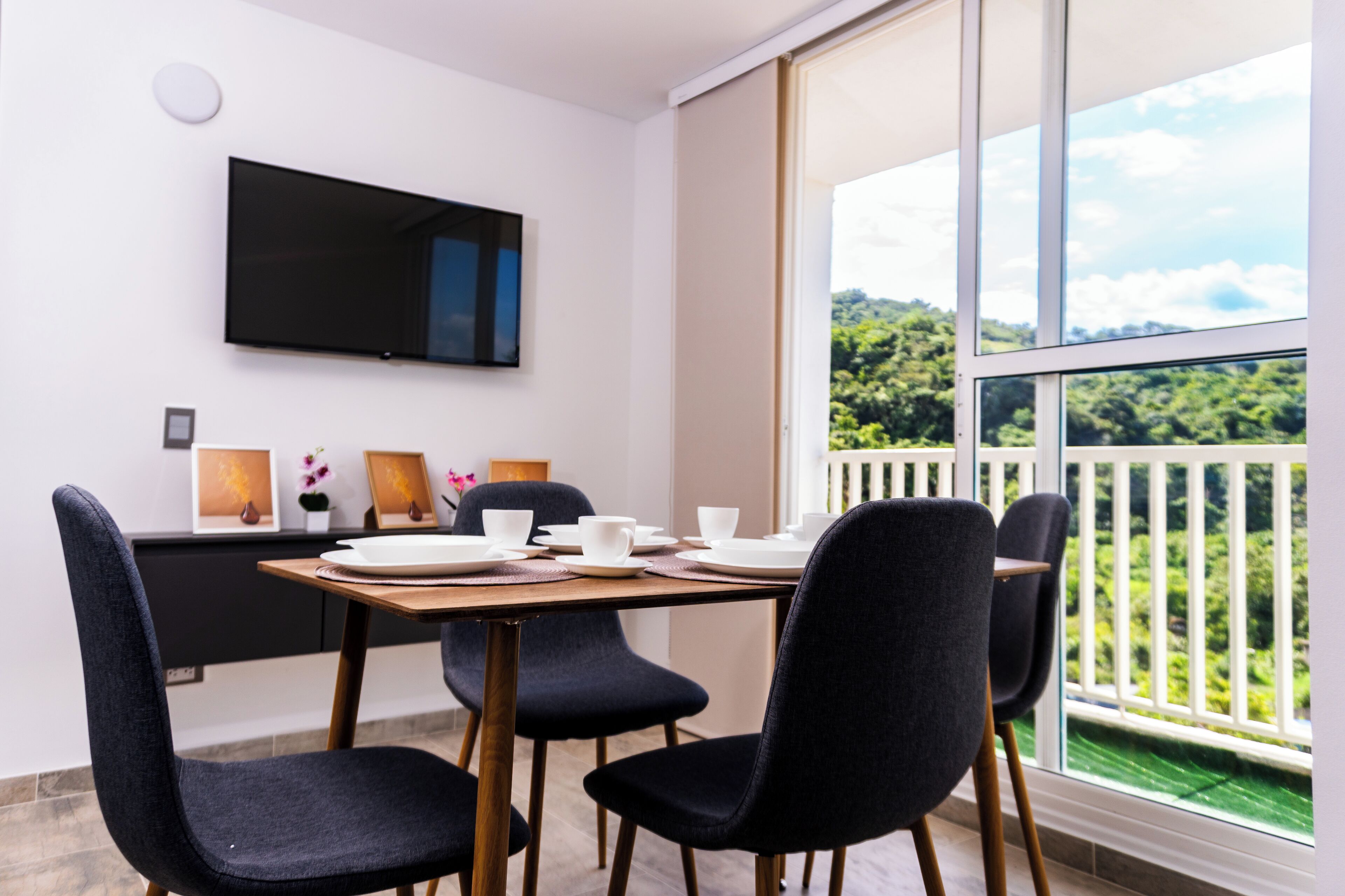Zona pranzo con tavolo apparecchiato per quattro persone, televisione a schermo piatto montata a parete e balcone con vista su area verde.
