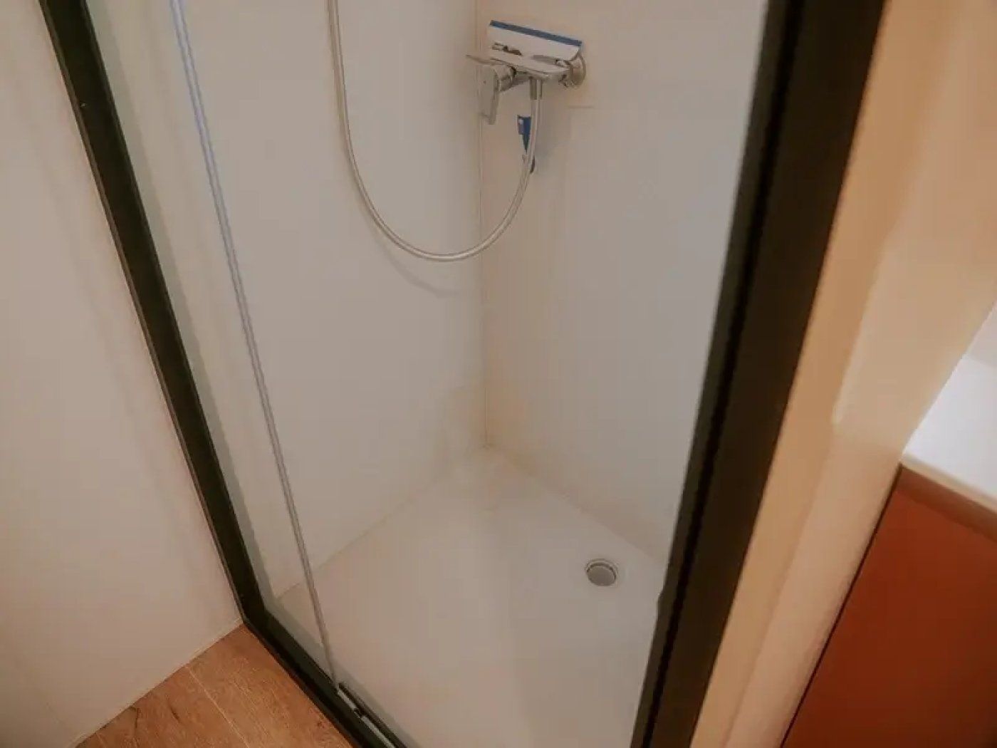 Área de chuveiro com divisória de vidro, piso de azulejo branco e ralo visível.