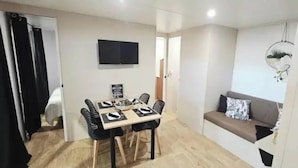 A compact living space with a dining area, a sofa, a TV mounted on the wall, and a hanging light fixture. - ZEcamping 3* piscine chauffée dog friendly animations locatif clim (Florac)