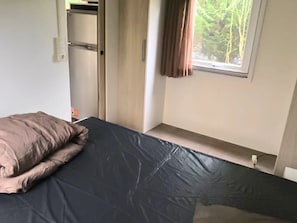 Un dormitorio con cama, ventana con cortinas, una puerta y un estante pequeño.