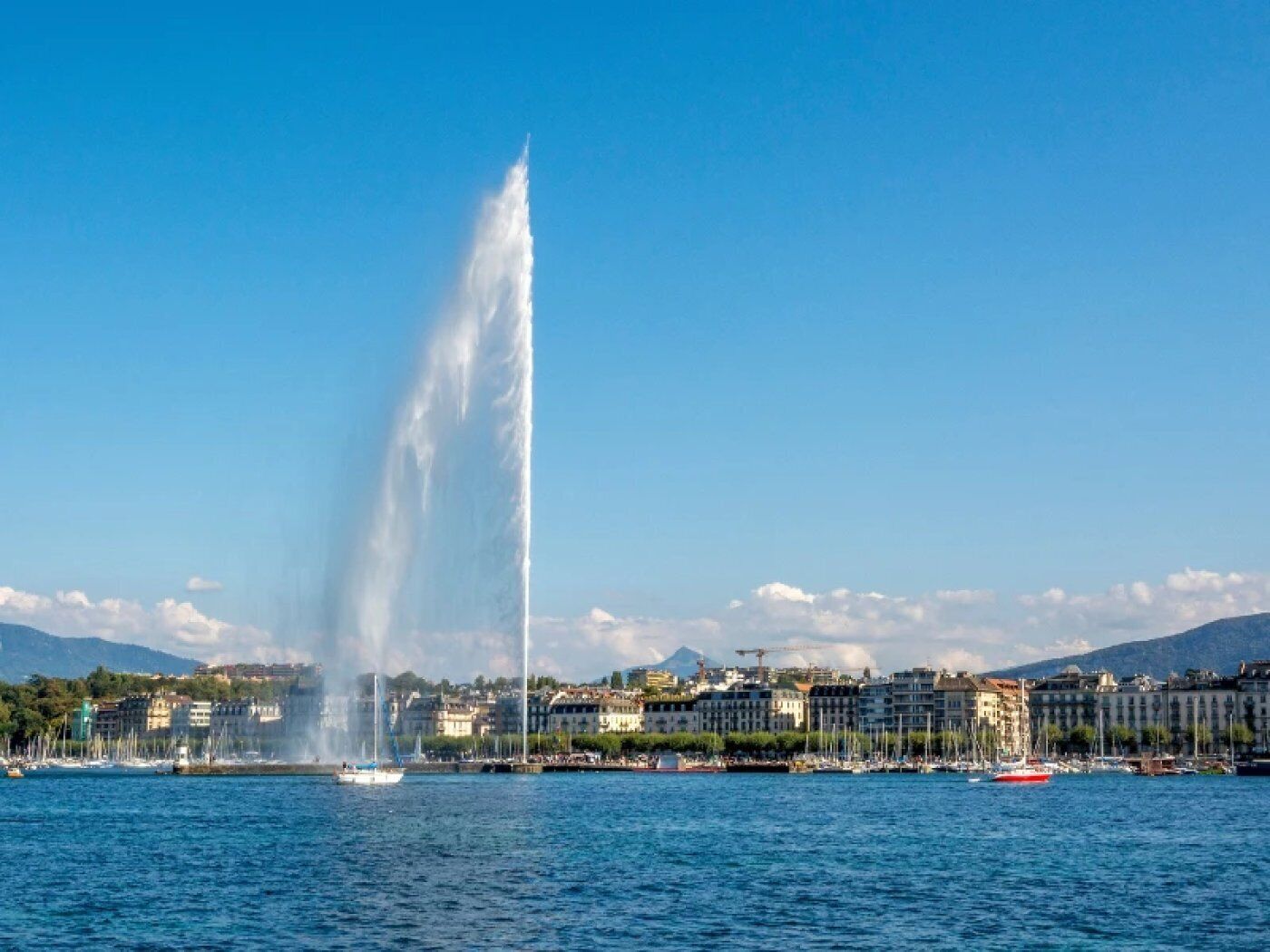 Une vue urbaine sur le front de mer, avec une fontaine imposante, des bateaux et des bâtiments qui bordent la rive.