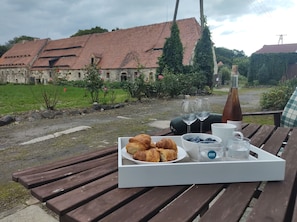 Ein Picknick mit Croissants, Blaubeeren und Weingläsern auf einem Holztisch mit Blick auf ein Steinhaus mit rotem Dach.