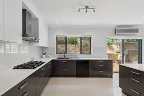 A modern kitchen with a central island, built-in appliances, and a large window providing a view of the outdoors. - Catos Escape Close to Rainforest and Scenic Lookouts (Mount Keira)