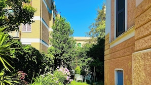 Un chemin de pierre mène à un bâtiment à la façade jaune, doté d’une fenêtre avec des volets.