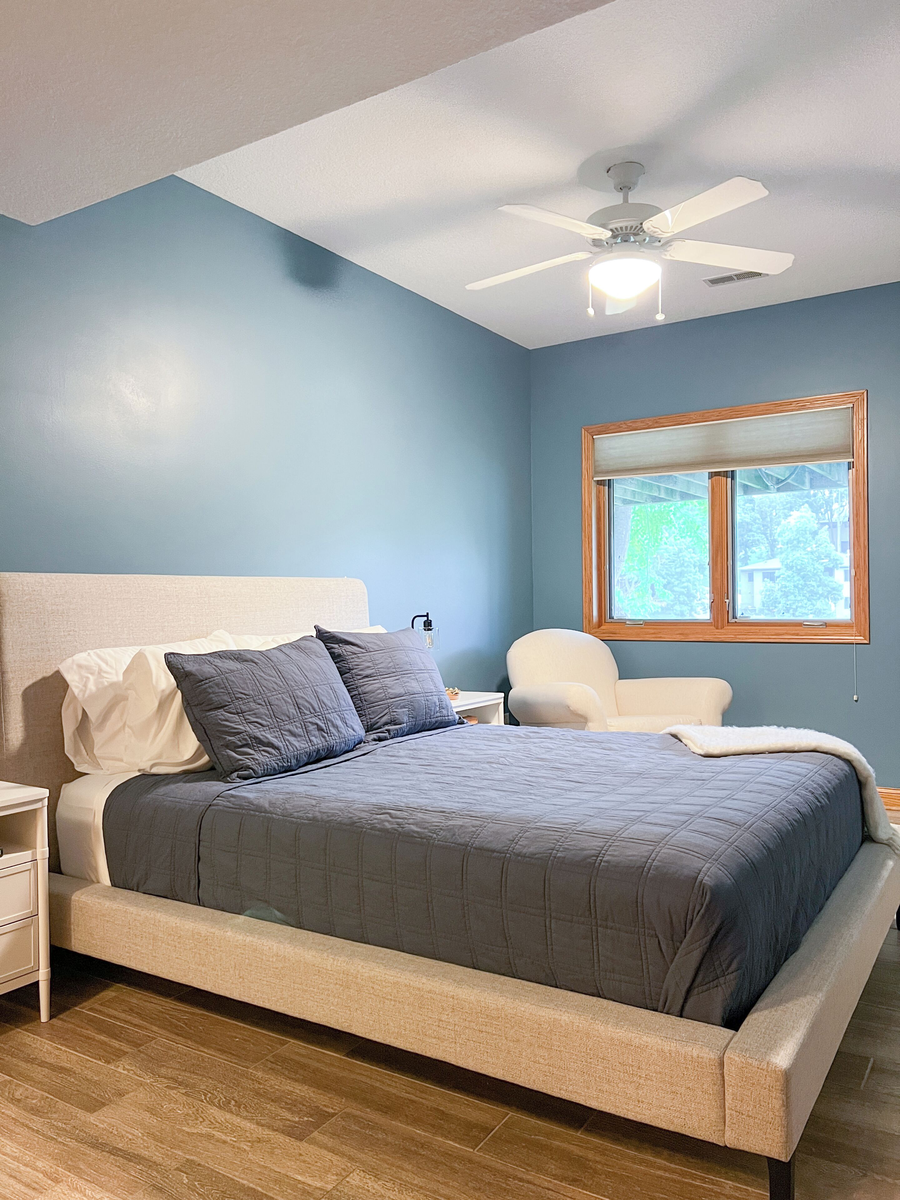 A bedroom with a bed, a nightstand, a chair, a ceiling fan, and a window.