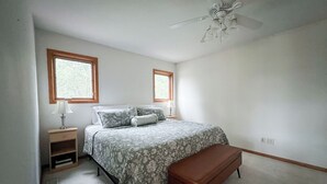 Une chambre à coucher comprenant un lit, deux lampes de chevet, une table de chevet, un banc, un ventilateur de plafond et deux fenêtres.