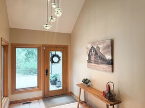Un couloir avec une porte en bois, un paillasson, un drapeau américain accroché au mur, une plante en pot, une petite table avec une plante et un tableau encadré représentant une grange.