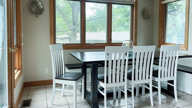Un coin repas avec une table et des chaises, de grandes fenêtres donnant sur la verdure et une feuille décorative accrochée au mur.