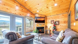 A cozy cabin interior with a fireplace, leather sofas, and a large window overlooking a scenic landscape. - Cozy Mountain Cabin w/ VIEWS | Hot Tub + Fireplace (Cosby)