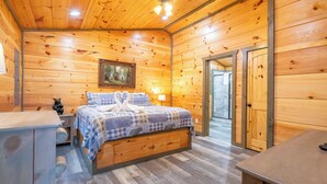 A wooden cabin bedroom with a bed, a nightstand, a lamp, and a painting on the wall. - Cozy Mountain Cabin w/ VIEWS | Hot Tub + Fireplace (Cosby)