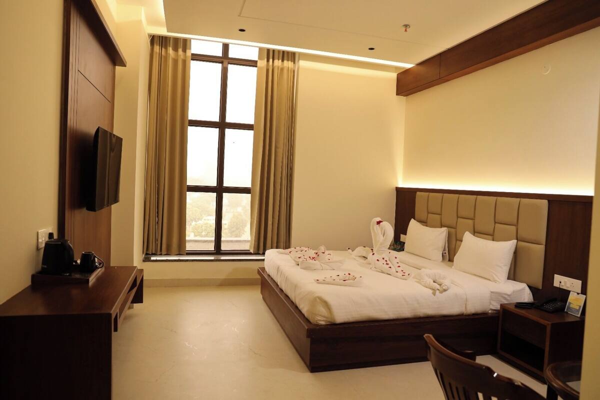 Ein Hotelzimmer mit einem großen Bett, einem an der Wand befestigten Fernseher, einem Schreibtisch mit Kaffeemaschine und einem Fenster mit Vorhängen.