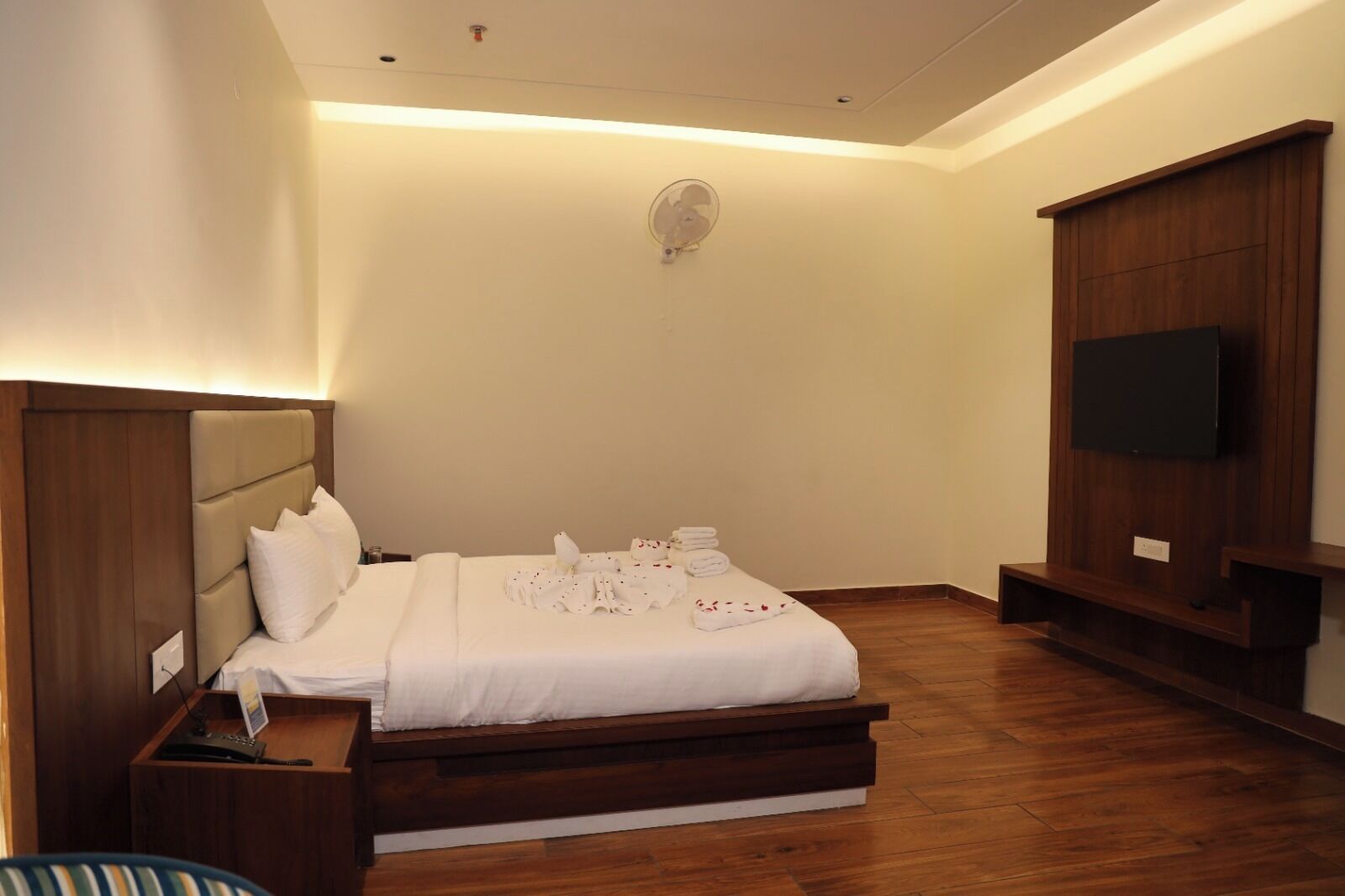 A bedroom with a bed, bedside table, TV, and a wall-mounted fan.