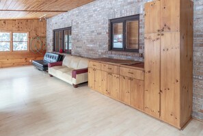 Ein Zimmer mit Backsteinwand, Holzboden, einem Ledersofa, einem Holzschrank und einem Fenster mit Blick auf Bäume.