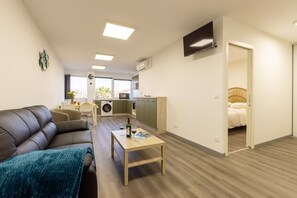 Ein modernes Wohnzimmer mit einer Couch, einem Couchtisch und einem Fernseher. Es gibt eine Küchenzeile mit einer Waschmaschine und einem Esstisch.