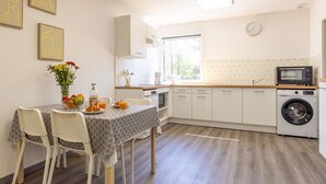 Een moderne keuken met witte kasten, een inbouwoven, magnetron en wasmachine. Er is een eettafel met stoelen en een bloevas.
